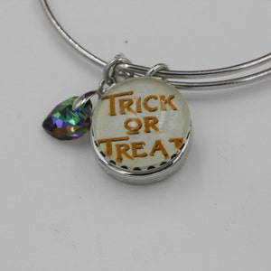 May include: Silver-toned bangle bracelet with a charm that says "Trick or Treat" in orange lettering. A colorful, iridescent crystal charm hangs next to the main charm. The bracelet is a festive accessory for Halloween.