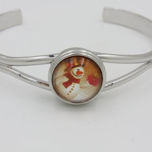 May include: A silver-toned cuff bracelet with a central circular design featuring a snowman wearing a red scarf and mittens. The snowman has a carrot nose and a top hat. The bracelet has a double-band design.