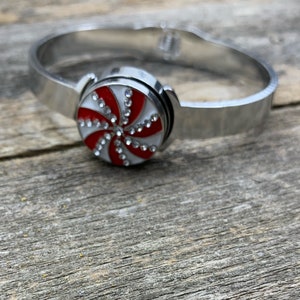 May include: A silver-toned metal bracelet with a decorative, round charm. The charm features a red and white peppermint swirl design, embellished with small, sparkling rhinestones. The bracelet is shown on a weathered wooden surface.