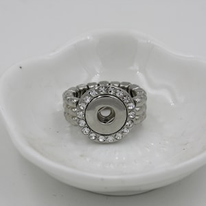 May include: A silver-toned ring with a large, round, central snap-on button, encircled by a row of clear, sparkling rhinestones. The band is a stretch style, and the ring sits in a white, petal-shaped dish.