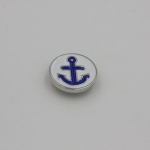 May include: A round, silver-toned button with a white enamel background. A blue anchor design is centered on the button. The button is likely used for clothing or accessories, and has a nautical theme.