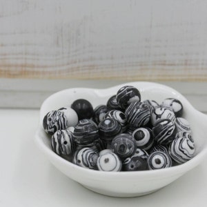 May include: A collection of black and white patterned beads in a white, heart-shaped dish. The beads have a marbled design, with varying shades of black and white. The dish is on a white surface, with a white wooden background.