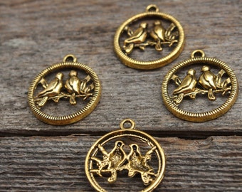 May include: Four antique gold-colored bird charms. Each charm features two birds perched on a branch within a circular frame. The charms have a loop at the top for attaching to jewelry or other crafts. The background is a weathered wooden surface.