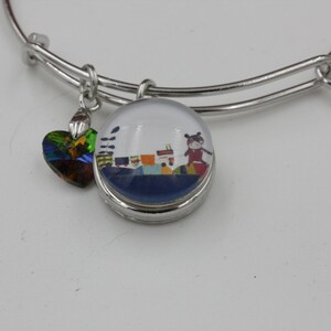 May include: Silver-toned bangle bracelet with two charms. One charm is a heart-shaped, iridescent crystal. The other is a round charm with a cartoon illustration of a person and colorful objects.