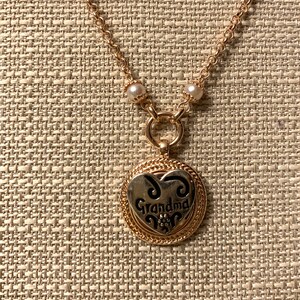 May include: A gold-toned necklace with a heart-shaped pendant that reads "Grandma" in a decorative font. The pendant has a silver-colored center and is surrounded by a gold-colored border. The necklace chain is also gold-toned, with small pearl accents.