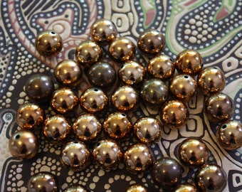 May include: A collection of shiny, metallic beads in shades of bronze and copper. The beads are spherical with a small hole at the top, and are scattered on a patterned fabric background. These beads are suitable for jewelry making and crafts.