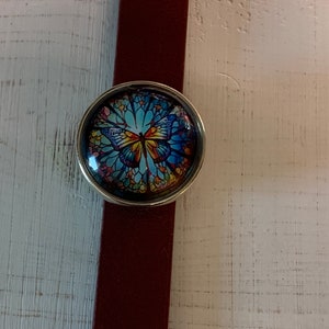 May include: A burgundy leather bracelet with a round, silver-toned button featuring a stained-glass style butterfly design in shades of blue, yellow, and orange. The bracelet is set against a white wooden surface.