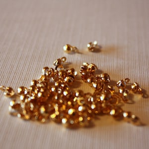 May include: A scattering of small, shiny, gold-coloured beads with attached loops, on a light-coloured fabric. The beads are round and appear to be made of metal, reflecting light. Suitable for jewellery making or craft projects.