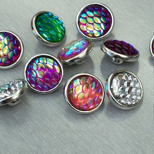 May include: A collection of decorative buttons with iridescent, scale-like patterns in various colors, including green, purple, and red. Each button has a silver-colored metal rim. The buttons are arranged on a light gray surface.