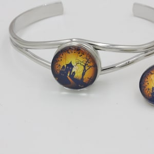 May include: A silver-toned cuff bracelet and matching stud earrings. Each piece features a round, Halloween-themed design with a haunted house, bare trees, and a dark road against a yellow and orange sky. The jewelry is set against a white background.