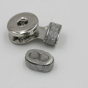 May include: A set of silver-toned magnetic clasps, featuring a circular top piece with a central hole and a curved connecting arm. The clasps have a textured, glittery finish on the edges of the oval-shaped bottom piece.