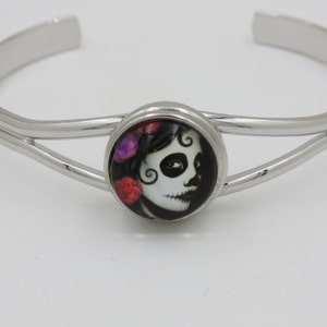 May include: A silver-toned cuff bracelet featuring a central circular design. The design showcases a Day of the Dead sugar skull with floral accents in red and purple. The bracelet has a double-band design.