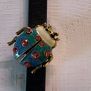 May include: A ladybug brooch with a black strap. The ladybug is blue and teal with pink accents and gold-colored trim. The brooch is attached to a black strap, possibly leather, against a white wooden background.