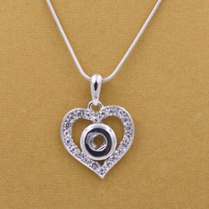 May include: Silver-toned necklace with a heart-shaped pendant. The pendant features a central snap-in button surrounded by a heart outline encrusted with small, clear crystals. The chain is a thin, snake chain style.