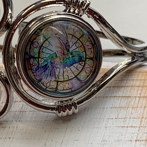 May include: Silver-toned metal cuff bracelet with a central round charm. The charm features a colorful stained-glass style image of a hummingbird in flight. The bracelet has decorative swirls and a hinged design.