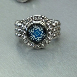 May include: Silver-toned ring with a round, blue and white snowflake design. The ring is embellished with clear, round rhinestones around the central design and along the band. The ring's band has a triple-row design.