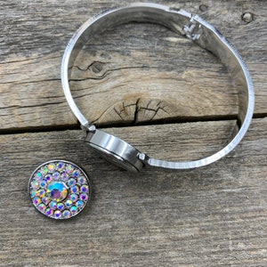 May include: A silver-toned hinged bracelet with a round watch face and a separate round charm. The charm is adorned with iridescent crystals in a circular pattern. The bracelet and charm are set against a weathered wooden surface.