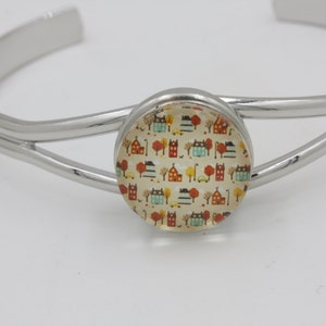 May include: A silver-toned cuff bracelet with a circular centerpiece. The centerpiece features a colorful pattern of houses, trees, and cars. The bracelet has a simple, elegant design, suitable for everyday wear.