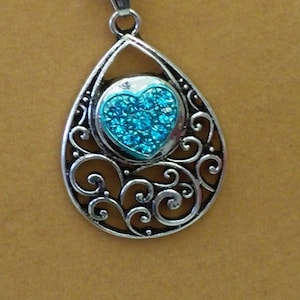 May include: Silver-toned pendant necklace with a teardrop-shaped frame. The pendant features a blue heart-shaped center with sparkling accents, surrounded by an ornate, swirling design. The necklace has a delicate ball chain.