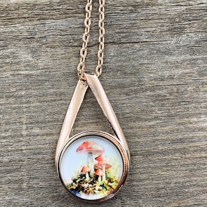 May include: Rose gold-toned pendant necklace featuring a circular charm with a mushroom illustration. The charm is set within a teardrop-shaped frame and hangs from a delicate chain. The mushrooms have red and white caps.