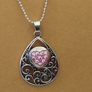 May include: Silver-toned pendant necklace featuring a teardrop-shaped frame with an openwork scroll design. The pendant holds a pink heart-shaped charm studded with smaller pink gems. The necklace has a delicate ball chain.