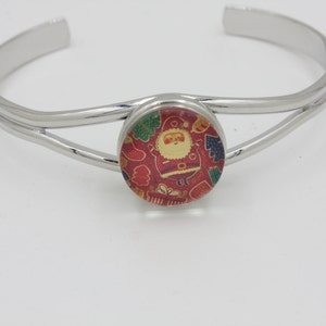May include: A silver-colored cuff bracelet with a festive Christmas-themed centerpiece. The round centerpiece features a Santa Claus design with red, green, and gold accents. The bracelet has a simple, elegant design.