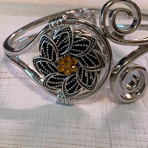 May include: Silver-toned cuff bracelet with a floral design. The bracelet features a central flower with black petals and a cluster of yellow gemstones. The design includes decorative swirls and a hinged opening.