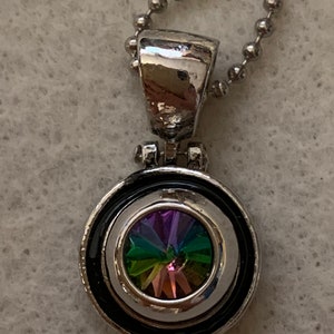May include: Silver-toned pendant necklace with a circular pendant featuring a vibrant, multi-colored crystal. The crystal displays hues of purple, green, and orange. The pendant is encircled by a black and silver frame, suspended from a ball chain.