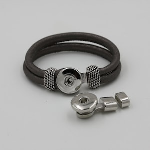 May include: A gray leather bracelet with a silver snap closure. The bracelet is double-wrapped and features a textured appearance. The snap closure is detached, showing the components. The bracelet is designed for wrist wear.