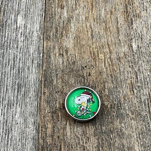 May include: A round, silver-toned snap button with a green background featuring a cartoon Snoopy character wearing a Santa hat and Christmas lights. The button is set against a weathered, gray wooden surface.