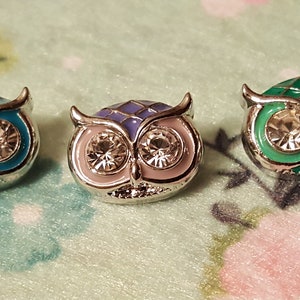 May include: Three owl-shaped charms with silver-colored metal frames. Each charm features a different color scheme: light blue, purple, and green. The owls have large, sparkling crystal eyes.