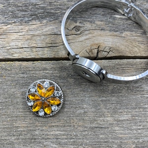 May include: A silver-toned hinged bracelet with a snap-on closure, next to a decorative snap featuring a yellow flower design with clear crystal accents. The jewelry is set against a weathered wood background.