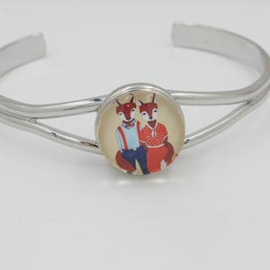 May include: Silver-tone cuff bracelet with a central circular charm. The charm features a whimsical illustration of two foxes, one in a red dress and the other in blue suspenders. The bracelet has a double-band design.