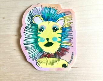 May include: A colorful sticker featuring a whimsical lion illustration. The lion has a yellow face, blue and green mane, and a red collar. The sticker has a pink and iridescent border and is placed on a light wood surface.