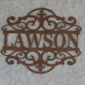 May include: A brown metal sign with the name "LAWSON" cut out in a decorative font. The sign has ornate scrollwork and is mounted on a textured, off-white wall. A decorative piece for a home.