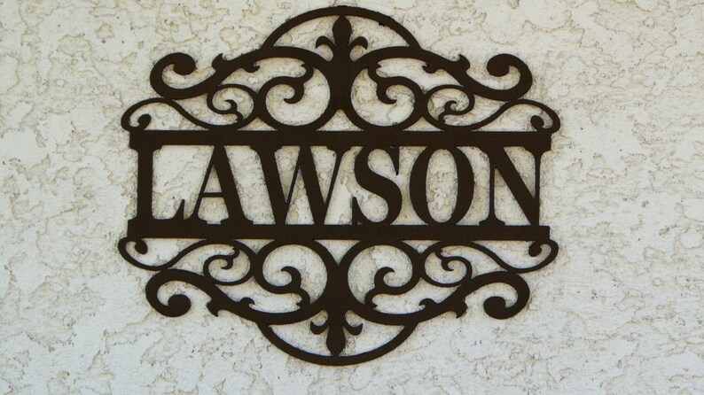 May include: A decorative metal sign with the name "LAWSON" cut out in bold letters. The sign has an ornate, scrollwork design around the name, with a dark brown finish. It is mounted on a textured, off-white wall.