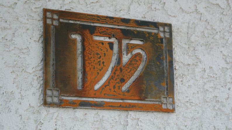 May include: A rectangular metal address plaque with the number 175 cut out. The plaque has a rusty orange and brown color scheme with decorative corner accents. It is mounted on a textured white wall.