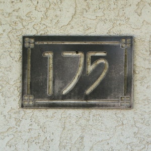 May include: A rectangular, dark metal address plaque with the number "175" in a stylized font. The plaque has a decorative border and is mounted on a textured, off-white stucco wall.