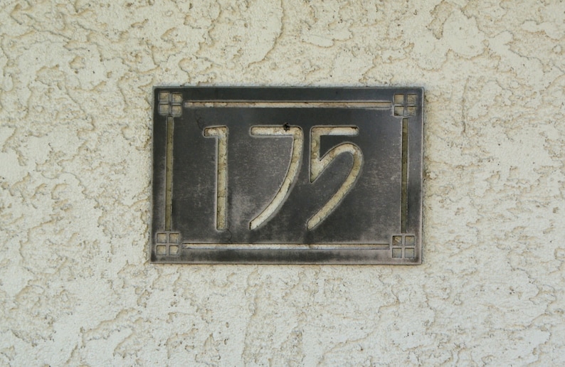 May include: A rectangular, dark metal address plaque with the number "175" in a stylized font. The plaque has a decorative border and is mounted on a textured, off-white stucco wall.