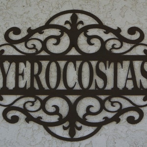 May include: A decorative metal sign with the name "YEROCOSTAS" in a stylized font. The sign is dark brown and features ornate scrollwork around the name, creating an elegant, custom look. The sign is mounted on a textured, light-colored wall.