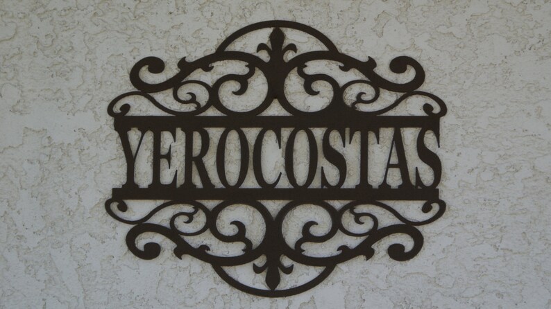 May include: A decorative metal sign with the name "YEROCOSTAS" in a stylized font. The sign is dark brown and features ornate scrollwork around the name, creating an elegant, custom look. The sign is mounted on a textured, light-colored wall.