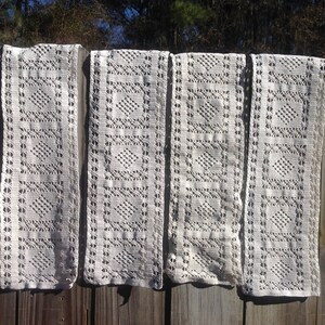 May include: Four white crocheted lace panels hanging on a wooden fence. The panels have a repeating square pattern.