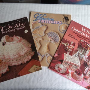 Könnte beinhalten: Drei Häkelanleitungshefte von Annie's Attic mit den Titeln Dolly Tote-Along Bassinet, Victorian Fridgies und Victorian Christmas Crochet. Die Umschläge zeigen Fotos von gehäkelten Gegenständen, darunter ein Stubenwagen, kleine dekorative Fächer und Geschenkboxen.