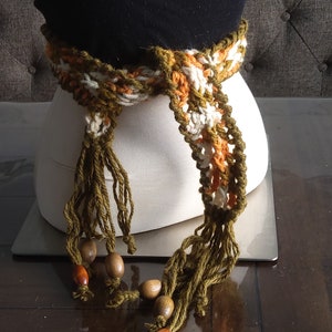 May include: A crocheted belt with brown, white, and orange stripes and long green tassels. The belt has wooden beads attached to the tassels.