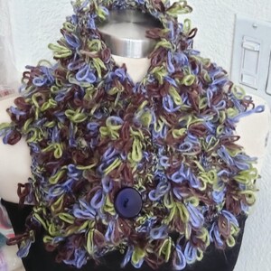 May include: A textured cowl scarf made of looped yarn in shades of brown, light green, and periwinkle blue. The scarf features a single large, round, dark purple button centered on the front, securing the looped knit fabric in a circular shape.