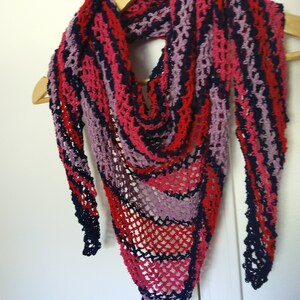 Pode incluir: Um xale de crochê feito à mão em forma triangular, exibido em um cabide de madeira. O xale apresenta uma paleta de cores vibrantes de vermelho, rosa, roxo e azul marinho, com uma trama aberta e rendada. É um acessório de moda.