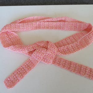 May include: A light pink crocheted scarf, tied in a knot. The scarf is made of a soft, textured yarn with subtle gold flecks. The scarf is lying on a white surface.
