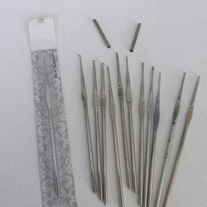 Puede incluir: Un conjunto de ganchillos plateados de varios tamaños, algunos con marcas visibles. Un ganchillo aún está en su embalaje original. También hay dos tubos de metal. Los artículos están dispuestos sobre una superficie blanca.