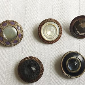 Puede incluir: Cinco botones circulares con diseños distintos. Uno presenta un patrón de diamantes morados con una incrustación nacarada central. Otro tiene un centro blanco con un borde texturizado. Un tercero es marrón oscuro con un centro facetado. Dos más son negros.