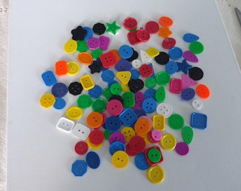 May include: A collection of colorful plastic buttons in various shapes, including circles, squares, stars, and triangles. The buttons are arranged in a random pattern on a white surface.